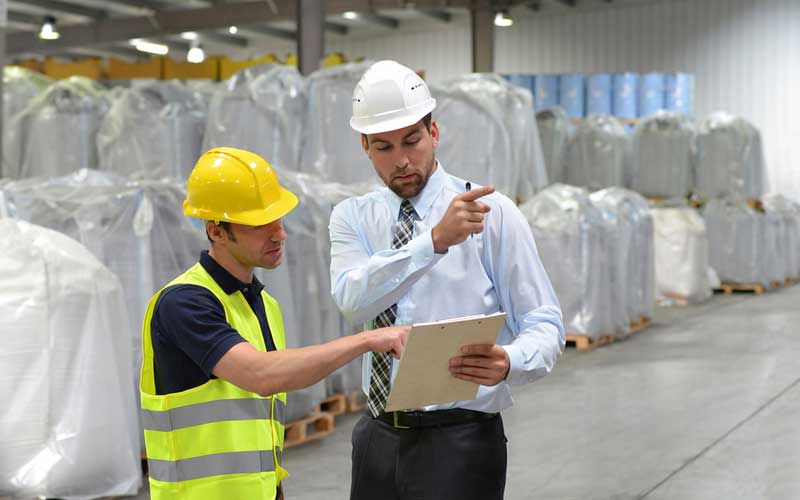 AW Associates warehouse workers checking clipboard wearing hard hats and hi vis jacket.